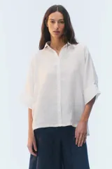 Camisa oversize blanca de lino, con corte cuadrado, mangas raglán cortas con puños anchos, aberturas laterales y cierre frontal de botones.