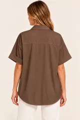 Camisa de algodón color chocolate, de diseño amplio y holgado, con cuello camisero, escote en V pronunciado con un botón decorativo dorado y mangas cortas con vuelta en los puños.