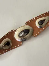 Cinto tipo faja de cuero color beige, con apliques metálicos plateados de forma ovalada y de gota, decorados con piedras oscuras y tachas metálicas a lo largo de toda la pieza.