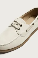 Mocasines tipo náutico de piel en color blanco, con suela plana de goma marrón, cordones y ojalillos metálicos laterales.