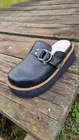 Zueco cerrado de cuero legítimo color negro, con plataforma y suela gruesa, adornado con una tira texturizada y anillas metálicas.