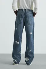 Jeans de corte relaxed fit en denim de algodón, color azul medio lavado. Presenta bordados florales en color blanco o crudo distribuidos en las piernas. Modelo de cinco bolsillos con cierre frontal de cremallera y botón.