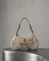 Cartera de mano de cuero vacuno con pelo largo en tono crudo y vainilla, con forma curva y solapa frontal con cierre magnético y aplique metálico con monograma. Cuenta con una correa ajustable corta para llevar al hombro o en la mano.