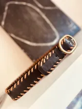 Cinturón de cuero negro con costuras decorativas color camel y hebilla ovalada de metal color bronce.