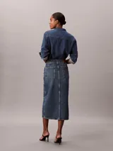 Falda midi de denim azul de talle alto, con diseño carpenter, cierre frontal de botón y cremallera, bolsillos y abertura central delantera.
