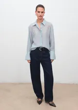 Blusa de tejido ligero color celeste con cuello camisero y cierre frontal de botones ocultos. Presenta mangas largas con puños anchos y detalles florales bordados en la parte superior delantera.