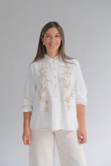 Blusa blanca de corte recto con cuello mao y volado pequeño, manga larga con puños elásticos y cierre frontal de botones. Presenta un delicado bordado floral en color beige en ambos lados del frente.