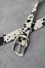 Cinturón ancho de cuero con textura de pelo y estampado animal print de manchas marrones y blancas. Cuenta con hebilla rectangular metálica con terminación envejecida y perforaciones regulables.