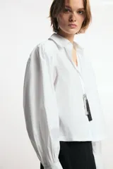 Camisa de cuello solapa y manga larga abullonada con puños abotonados. Presenta un diseño de corte recto con cierre frontal oculto por solapa.