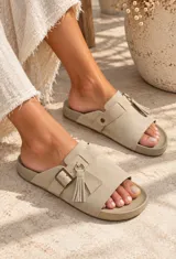 Sandalias planas color beige, fabricadas en gamuza, con tira ancha en el empeine, hebilla metálica de ajuste y detalle de flecos colgantes con argolla.