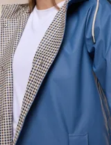Pilot engomado impermeable color azul, de corte recto estilo trench con acabado resistente al agua. Cuenta con capucha integrada, cierre frontal con broches a presión, bolsillos laterales con ojal y forro interior de tela estampada a cuadros visible en la solapa y los puños.