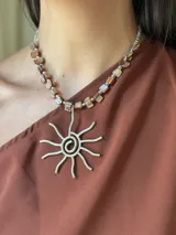 Collar corto con dije de sol estilizado en metal plateado y cadena de cuentas cuadradas de nácar en tonos marrones y beige, intercaladas con mostacillas plateadas.