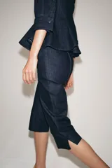 Pantalón capri de tiro medio confeccionado en denim de algodón. Presenta bolsillos de vivo en el delantero, cierre frontal con cremallera, botón y ganchos metálicos, y aberturas laterales en el bajo.