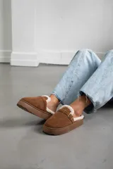 Zueco de gamuza color marrón, con interior forrado en corderito sintético, suela de plataforma plana y detalle de tira con hebilla metálica en el empeine.