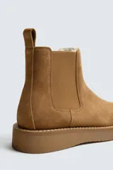 Bota estilo Chelsea de hombre, confeccionada en piel con acabado de serraje en color tostado. Presenta elásticos laterales a tono, horma redonda y suela plana del mismo color.