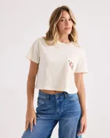 Remera corta de corte boxy en color blanco vintage, con mangas cortas, cuello redondo y un bolsillo aplicado en el pecho que cuenta con un pequeño estampado floral y de serpiente.