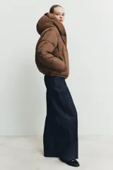 Campera puffer acolchada de color negro, con cuello envolvente, capucha integrada, manga larga, bolsillos laterales ocultos y bajo con elástico. Cierre frontal con botones a presión ocultos por solapa.