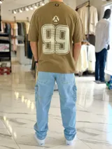Remera regular color off-white, con diseño gráfico en la espalda que incluye el número 99 en gran tamaño y detalles en negro. Cuenta con ribetes negros en las costuras de los hombros y mangas.