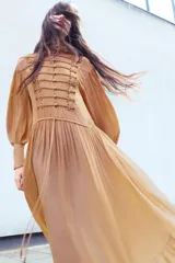 Vestido largo de viscosa en color tostado, con cuello subido y manga larga terminada en puño. Cuenta con cintura ajustable con lazadas, forro interior, bajo evasé y cierre frontal con alamares y botones.