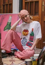 Remera blanca de corte recto con cuello redondo y estampado de objetos cotidianos: tomates, botella de vino, jarrón y latas de sardinas.