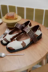 Zapatos tipo Mary Jane con punta cuadrada, confeccionados en cuero con estampado de vaca (animal print) y tira ajustable con hebilla metálica en color marrón.