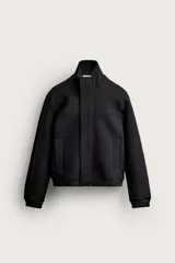 Cazadora de corte boxy fit en tejido compacto con elasticidad, color negro. Presenta cuello subido, manga larga con puños elásticos, bolsillos frontales de vivo y cierre frontal con cremallera oculta por solapa con botones a presión.