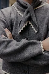 Chaqueta de punto gris oscuro, corte cropped y manga larga, con cuello redondo que incorpora una bandana anudada al frente. Los bordes del cuello, puños y ruedo están terminados con una puntilla de encaje color beige claro. Cierre frontal con botones oscuros.