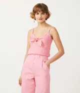 Blusa musculosa de color rosado confeccionada en tejido laise de algodón. Presenta un escote en V con un lazo decorativo en el frente, tirantes finos y detalle de lastex en la espalda para un mejor ajuste.