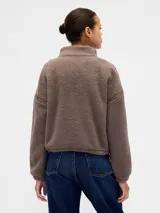 Buzo de polar tipo sherpa color marrón, con cuello alto y cierre frontal parcial. Presenta el logo de la marca bordado en el pecho y detalles de costuras reforzadas en hombros y mangas.