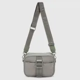 Cartera tipo bandolera de diseño compacto en color gris, confeccionada en material sintético con acabado satinado. Presenta un compartimento principal con cierre, un bolsillo frontal con solapa y hebilla decorativa, y una correa larga ajustable con el logo de la marca estampado.