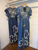 Vestido largo de denim azul con escote en V, mangas cortas con volados y cordón ajustable con borlas en la cintura. Presenta bordados detallados en el pecho y apliques grandes de flores con flecos en la parte inferior de la falda.