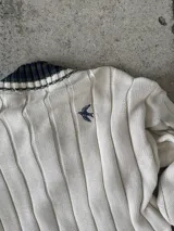 Buzo tejido de punto con escote en V, manga larga y diseño holgado. Presenta un patrón de rayas verticales en relieve y detalles de franjas en contraste en el cuello, puños y ruedo. Incluye un pequeño bordado en el pecho.