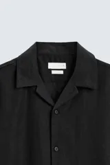Camisa de lino color negro con calce relajado, cuello bowling y manga corta. Presenta cierre frontal con botones a tono y aberturas laterales en el bajo.