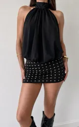 Vestido corto negro con escote halter drapeado y falda ajustada con detalle de tachas doradas cuadradas.