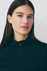 Campera deportiva negra híbrida con cuerpo acolchado y mangas de tejido elástico. Presenta cuello alto, cierre frontal con protección para el mentón, bolsillos laterales y bolsillos interiores.