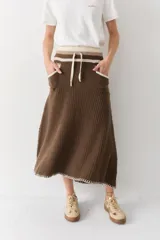 Falda midi de lana en color marrón, con diseño acampanado y tejido acanalado. Presenta una cintura elástica con cordón de ajuste, detalles de rayas en tonos beige y blanco en la parte superior, y costuras decorativas en el ruedo.