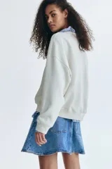 Buzo de cuello redondo en color beige, con hombros caídos y ajuste holgado. Presenta una letra A grande en relieve con textura afelpada en el centro del pecho y un pequeño bordado debajo. Cuenta con puños y cintura acanalados.