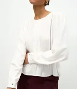Blusa tipo bata de color blanco, confeccionada en viscosa, con cuello redondo, mangas largas con puños abotonados, detalles de entremedios verticales y un recorte con volado en el bajo.