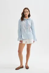 Camisa de algodón con detalles de broderie, volados y cuello a la base. Presenta diseño holgado, cierre frontal con botones y mangas largas con puños fruncidos.