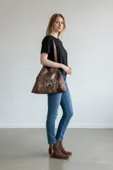 Cartera de cuero natural en color marrón jaspeado, tipo tote bag, con diseño amplio y asas dobles para el hombro. Presenta intervención artística pintada a mano en el frente con detalles metalizados y geométricos, y dije metálico colgante.