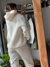 Buzo tipo hoodie de corderito color manteca, con capucha y cordones ajustables. Presenta un diseño abstracto frontal con apliques de textura suave en tonos rosa viejo y marrón.