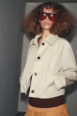 Trench corta color beige con cuello solapa, manga larga con trabilla y botón en los puños, bolsillos de vivo en el delantero y cierre frontal con botones oscuros.
