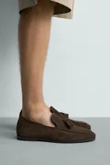 Mocasines de vestir para hombre confeccionados en piel con acabado en gamuza de color marrón, con detalle de borlas en el empeine y suela plana con ligero tacón.