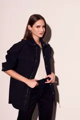 Sobrecamisa negra de algodón con corte oversize, cuello clásico, cierre frontal con botones a presión, dos bolsillos de parche en el pecho y apliques metálicos en cuello y solapa.