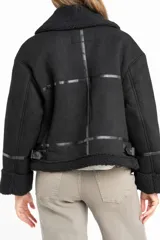 Campera tipo gamulán de corte oversize, color negro, con interior y detalles en cuello y puños de corderito sintético. Presenta cierre frontal y correas decorativas en el cuello.