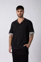 Camisa tejida negra de manga corta con cuello bowling y textura trenzada vertical. Los puños tienen un detalle de rib con dos franjas blancas.