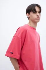 Remera de algodón teñida en prenda, color coral/rosado, con calce boxy y estampado en la espalda con texto en tipografía cursiva y líneas de texto más pequeñas.