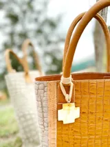 Bolso matero de cuero color marrón con textura croco y detalles en cuero color taupe. Tiene doble asa corta de cuero y un dije colgante en forma de cruz.