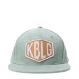 Gorro tipo cap de pana color verde agua, con parche hexagonal frontal en color tostado y letras blancas con las siglas KBLG.