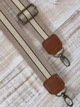 Correa de repuesto para cartera, de tela color beige con rayas blancas y negras. Incluye herrajes metálicos color bronce y apliques de cuero sintético color marrón.